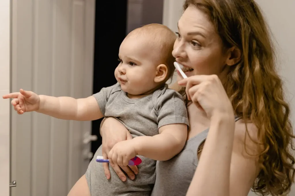 A mother with long brown hair stands in a bathroom, looking into a mirror and brushing her teeth, while holding her baby dressed in a gray onesie. The baby is pointing off-camera and holds a small toothbrush, learning about daily routines.