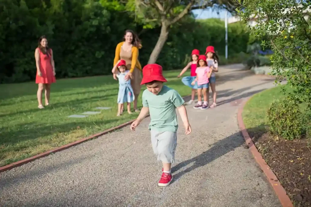 A group of young children and their adult caregivers are enjoying a sunny day outdoors in a park or garden. A toddler boy in a red hat is walking or running down a gravel path toward the foreground, while a line of other children and two adults watch from the background.