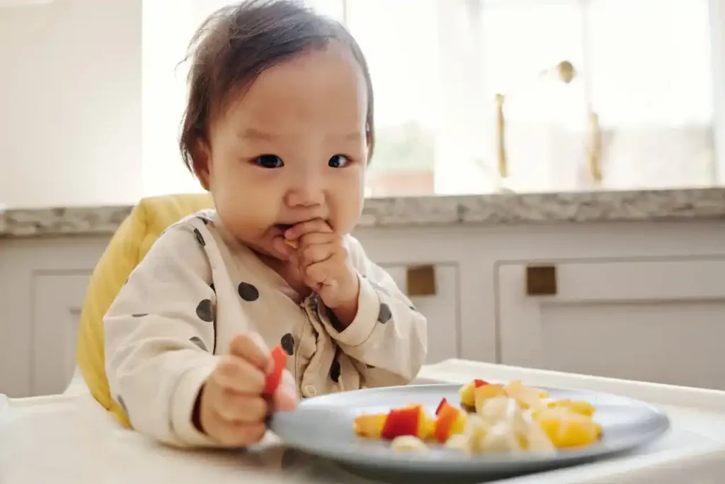 Baby in a high chair self-feeding soft, diced fruit from a plate—Solid Foods for Babies with bite-size, easy-to-grip pieces.