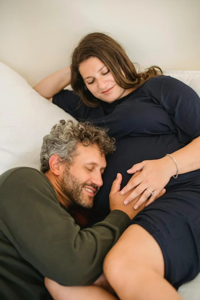 A pregnant woman wearing a dark dress is lying comfortably on a white bed or couch while her partner, a man with curly grey hair and a beard, is leaning in to affectionately place his ear against her large belly. They are both smiling with their eyes closed, showing a moment of calm connection and mutual support.