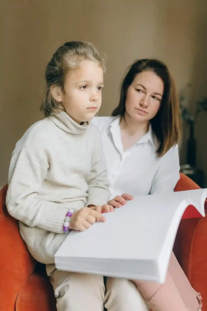 A young girl in a cream-colored turtleneck sweater is sitting in an orange chair, looking thoughtfully to the side while holding a large, thick white book or binder. Her mother sits behind her, looking on with a supportive, focused expression. The scene suggests a parent and child engaging with a structured tool, perhaps for supporting children with special needs in everyday routines.