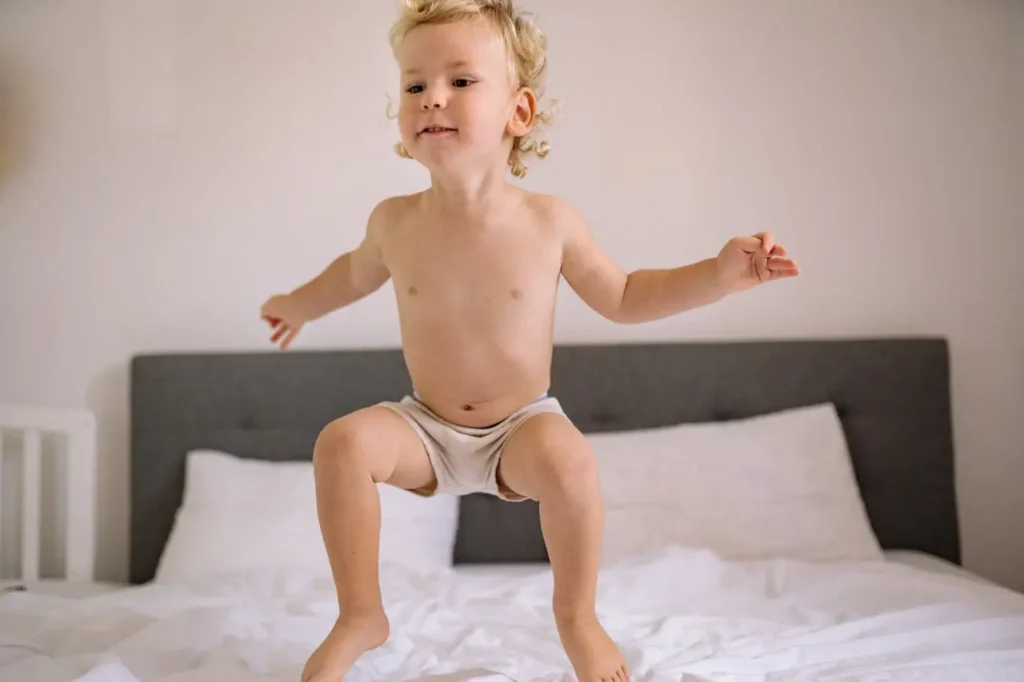 A blonde, curly-haired toddler wearing only light-colored underwear is captured mid-air, jumping enthusiastically on a white bed with a simple gray headboard. The image conveys active gross motor development and playful energy.