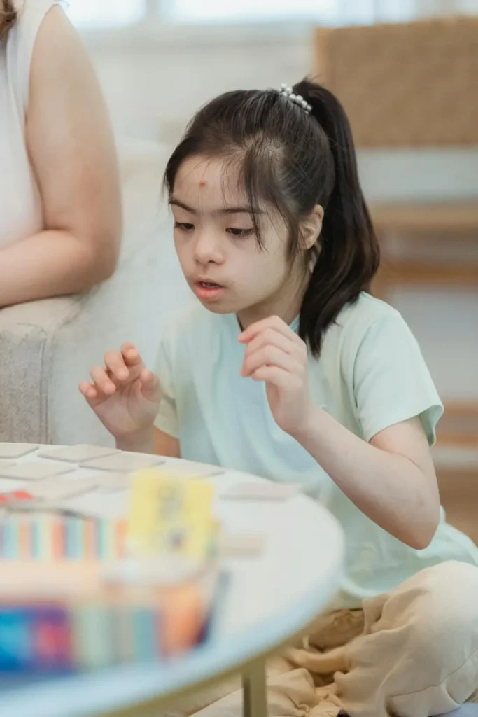 A young girl, who appears to have Down syndrome, with dark hair tied in a ponytail, is sitting on a floor or low cushion, deeply focused on a set of small, square cards or tiles spread out on a low, light-colored table. She is wearing a light green T-shirt and has her hands raised slightly above the cards as if contemplating her next move in a game or activity.