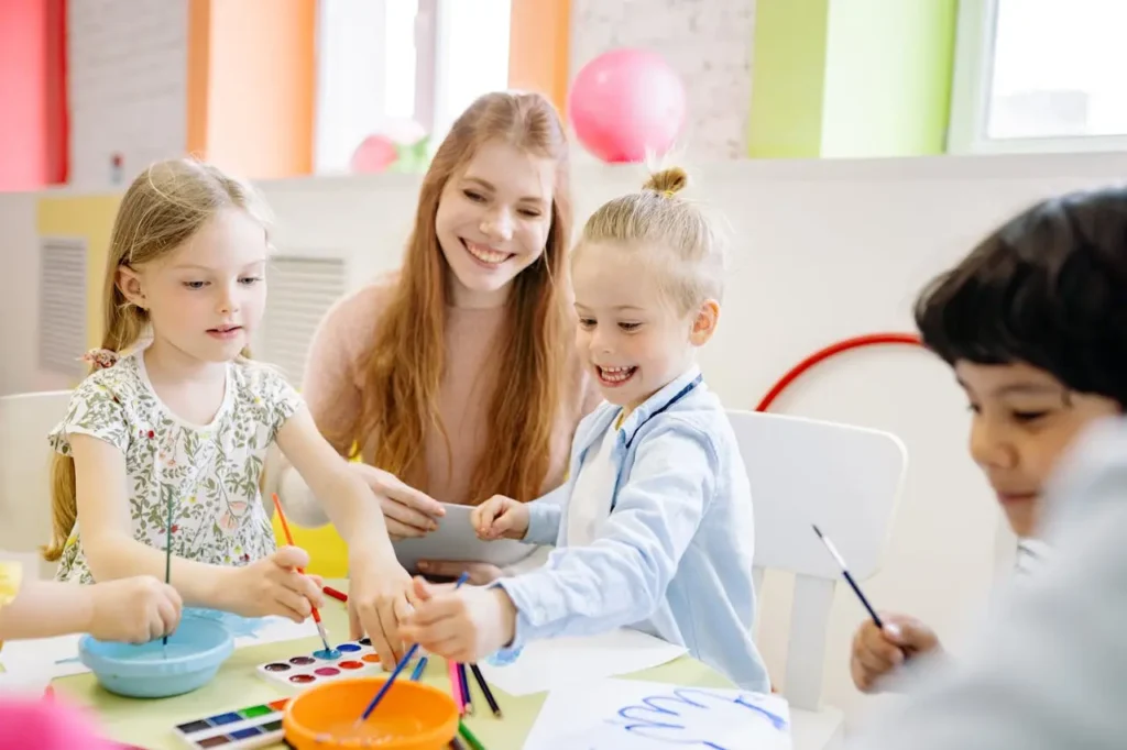 A smiling female teacher with long red hair is assisting three young children sitting at a table during an art activity involving paints, brushes, and paper. The children are happily engaged, but the image highlights the necessary close interaction between a caregiver and a child.