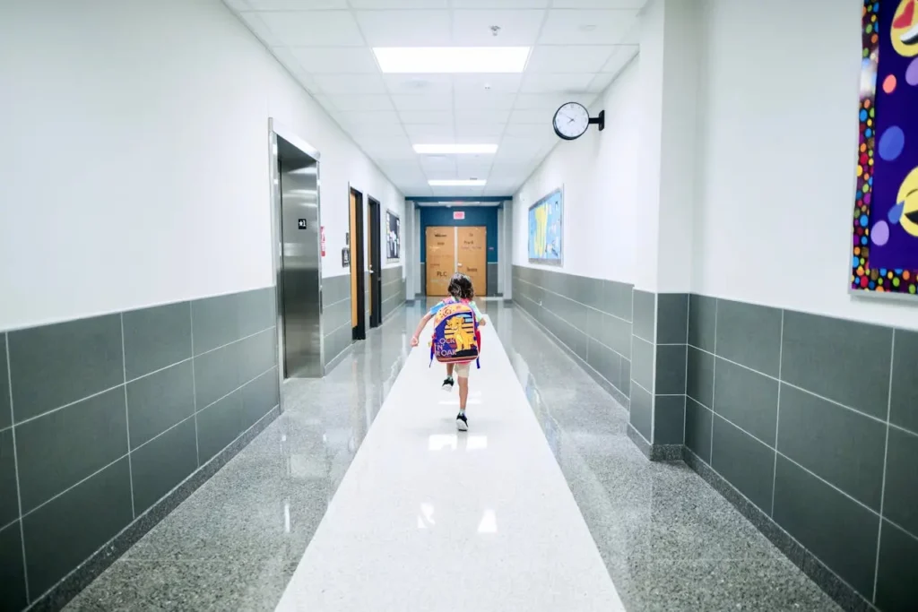 A small child wearing a colorful backpack runs down a bright school hallway toward classroom doors.