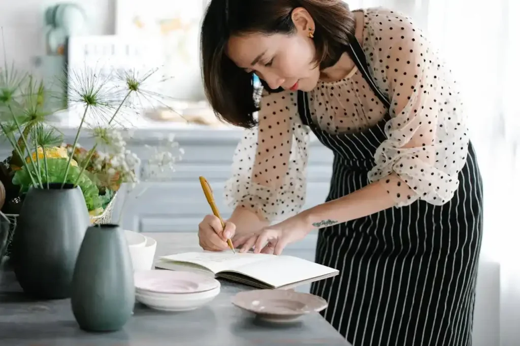 A woman with dark hair in a polka-dot apron is leaning over a table, thoughtfully writing or planning a list in a small open notebook using a gold pen. She is surrounded by vases and flowers, suggesting a home setting.