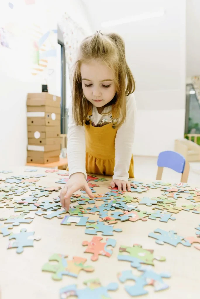 A vertical shot of a young girl with blonde pigtails, wearing a yellow dress, standing at a table and deeply concentrating on assembling a large pile of colorful jigsaw puzzle pieces.