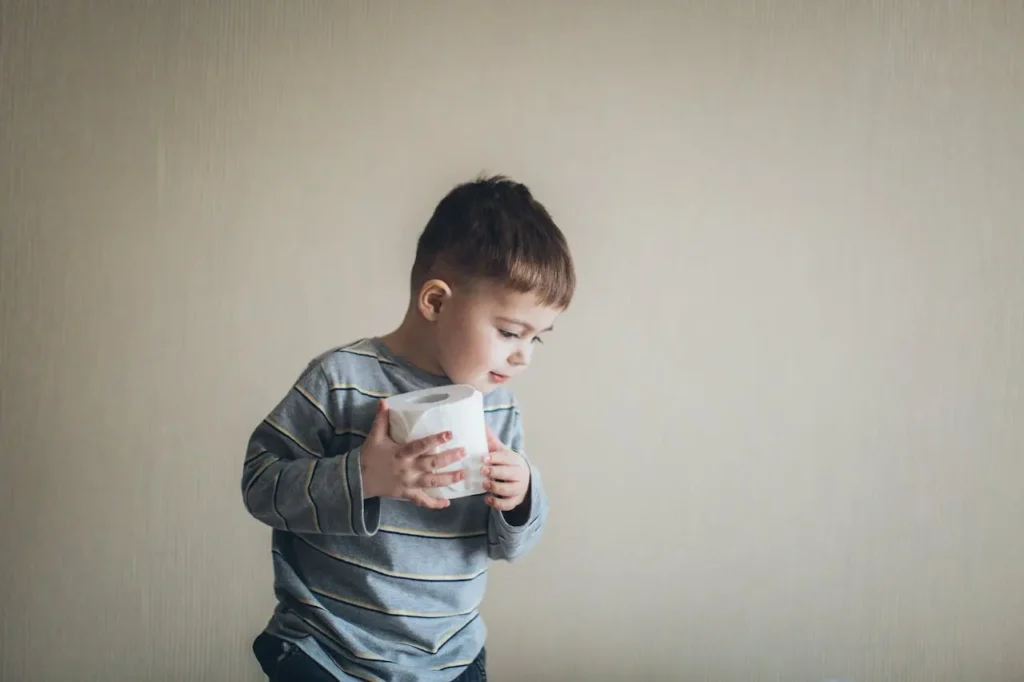 A young boy in a striped shirt, approximately 2-3 years old, is standing against a plain beige wall, intently holding and looking at a full roll of toilet paper, symbolizing a step in a Potty Training Plan.
