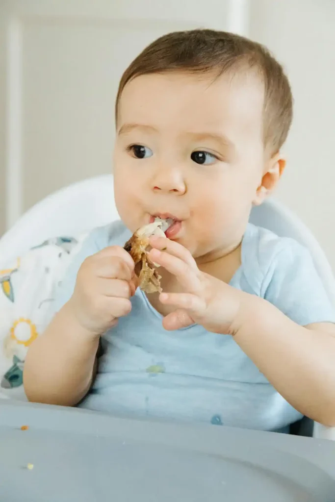 A young toddler sitting at a small table, happily picking up and eating colorful, chopped fruits and vegetables, such as blueberries and sliced carrots, from a divided plate.