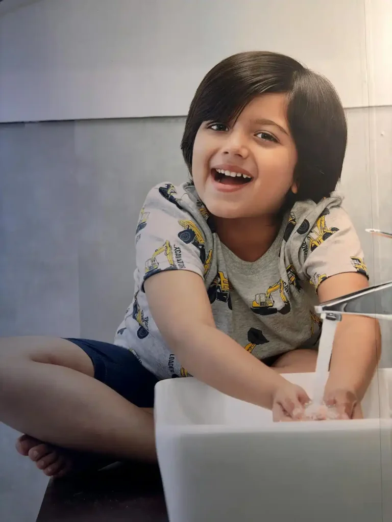 A young child with dark hair and a wide smile is sitting on the edge of a counter or sink, actively washing their hands under running water in a white sink basin. The child is wearing a gray t-shirt with construction truck prints.