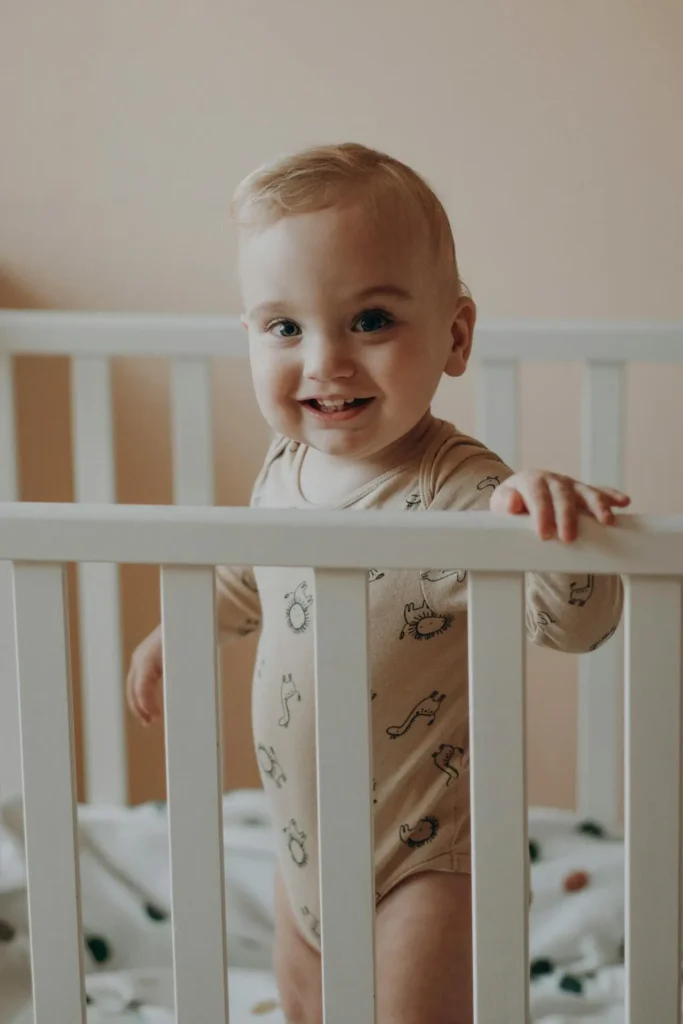 A blonde toddler boy wearing a light-colored onesie with small animal prints is standing up inside a white wooden crib, holding onto the top rail and smiling happily at the camera.