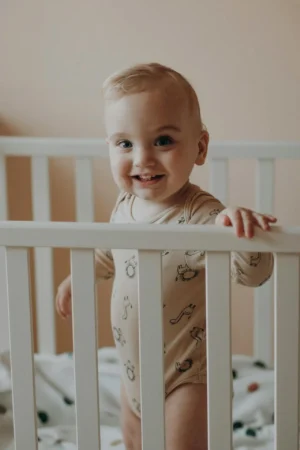 A blonde toddler boy wearing a light-colored onesie with small animal prints is standing up inside a white wooden crib, holding onto the top rail and smiling happily at the camera.