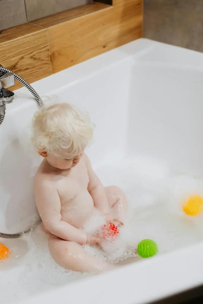 A fair-skinned toddler with very light, curly blonde hair is sitting in a white bathtub filled with bubbles, playing with a small red toy. Orange and green bath toys float nearby.