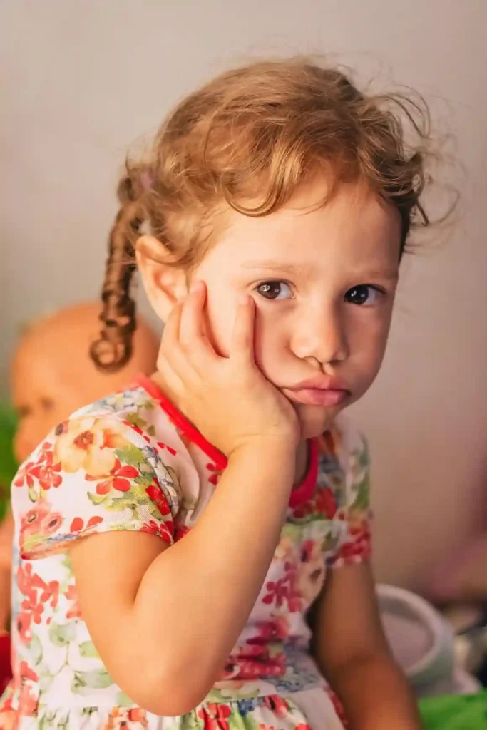 A close-up portrait of a toddler girl with curly brown pigtails, wearing a floral dress, resting her hand on her cheek and looking directly at the camera with a visibly sad, serious, or distressed expression.