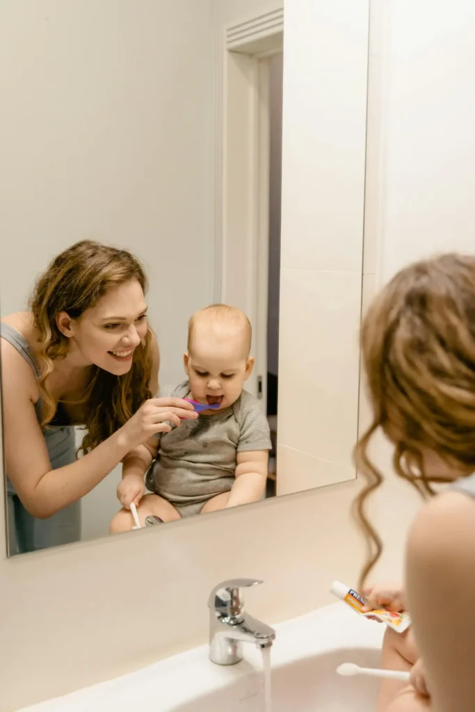 A mother smiling while helping a baby or young toddler seated on the bathroom counter brush their teeth with a small purple toothbrush, visible in the mirror's reflection.