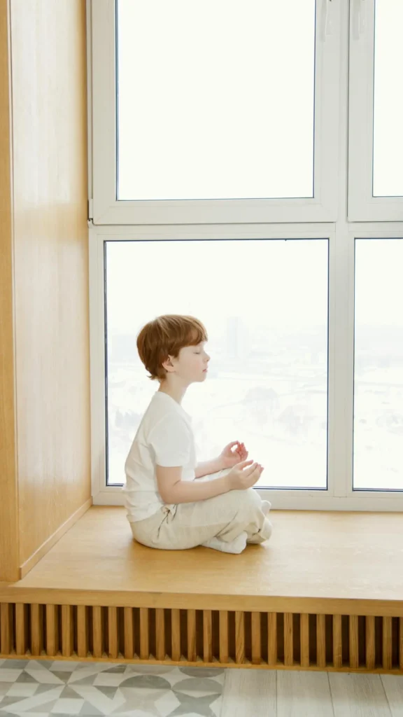 A young boy with reddish-brown hair, wearing a white t-shirt, sits cross-legged in the lotus position on a wooden windowsill ledge with his eyes closed, practicing a relaxation technique.