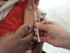 A close-up shot showing the arms and hands of two adults and a child. One adult is holding the child's upper arm steady, while a second adult is administering an injection (a vaccine) into the child's arm using a syringe.