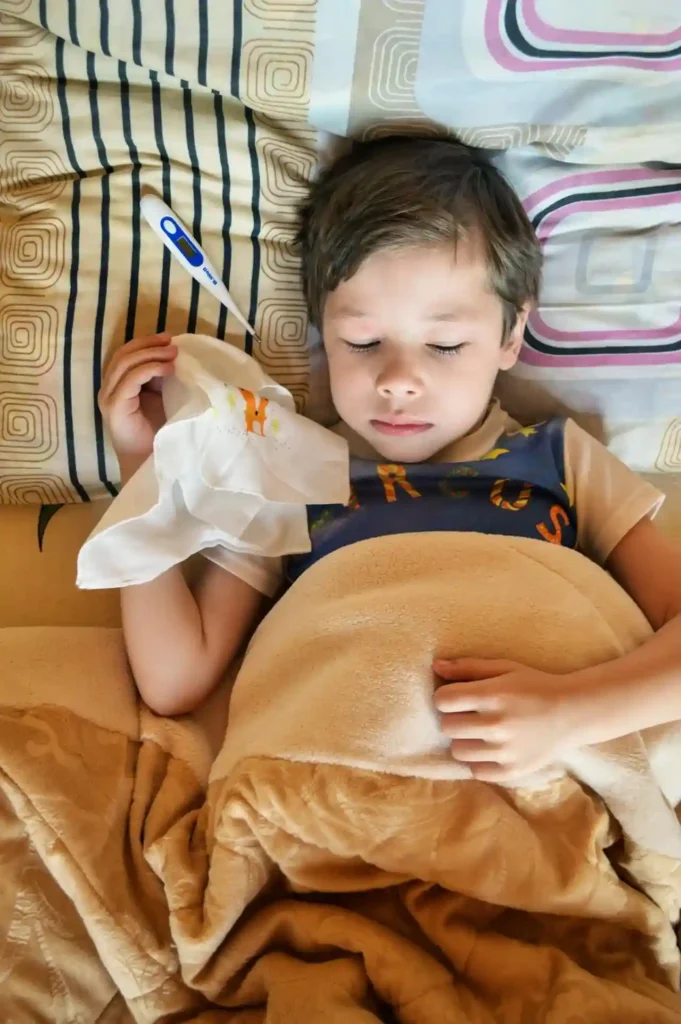 A young boy is lying in bed, covered with a blanket, with his eyes closed. He is holding a digital thermometer in his hand near his chest, indicating that he is ill, possibly with one of the common childhood illnesses.