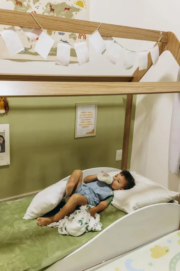 A toddler boy with dark hair is lying down comfortably in a white wooden house-frame bed, propped on pillows, drinking from a bottle. The child is wearing blue denim overalls and a green shirt, and the room is decorated with a world map and a banner.