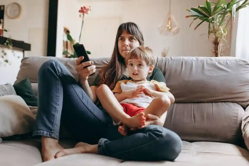 A mother with long dark hair, wearing jeans and a t-shirt, is sitting barefoot on a gray sofa, holding her young son on her lap while looking down at a smartphone in her hand. The boy is looking directly ahead.