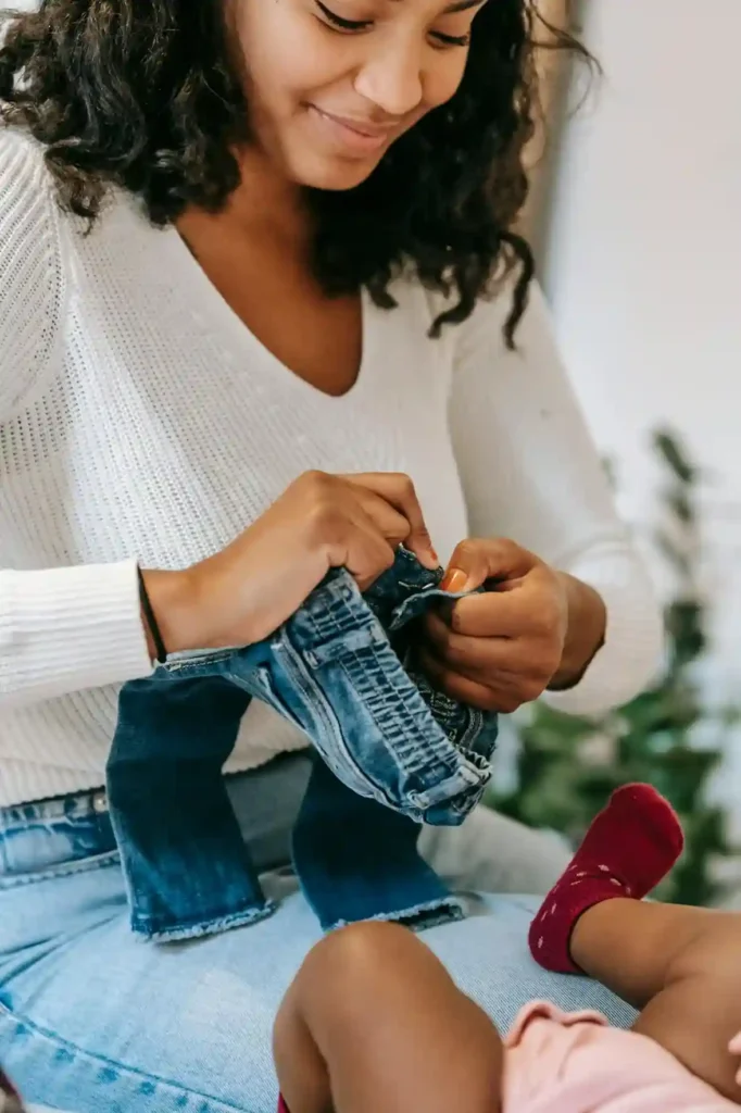 A close-up of a smiling mother with curly dark hair, wearing a white sweater, carefully pulling a pair of denim jeans onto the legs of a baby, who is wearing a red sock on one foot.