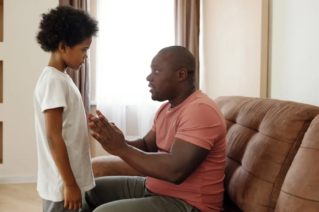 A man sitting on a brown couch in a living room is speaking seriously to his young son who is standing in front of him. The father's hands are clasped, and the boy's face shows an intense, serious expression, depicting a moment of setting Family Rules That Kids Understand.