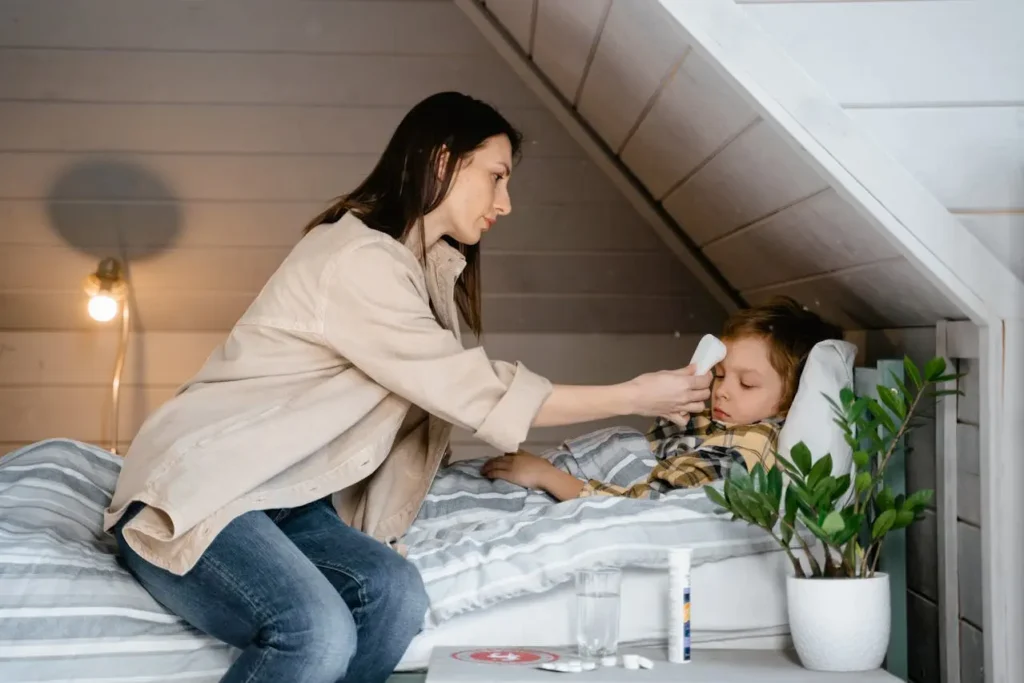 A mother in a beige jacket and jeans sits on the edge of a bed, gently holding a digital thermometer or cold compress to the forehead of her young son who is resting under a striped blanket, clearly depicting care for a child with a fever in toddlers.