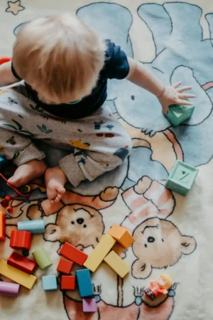 A high-angle shot of a blonde toddler sitting on a patterned play mat, reaching out to grasp or move a small green cube. Many brightly colored wooden blocks are scattered around the child, indicating play that targets fine motor skills.