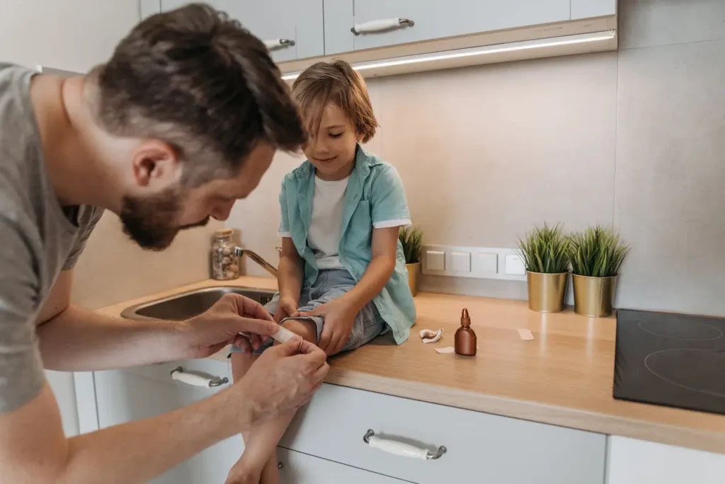A man with a beard, wearing a white t-shirt and light blue shirt, is kneeling to apply a small bandage to the knee of his young son, who is sitting on a kitchen counter. The boy is smiling, showing comfort and trust during the injury care.