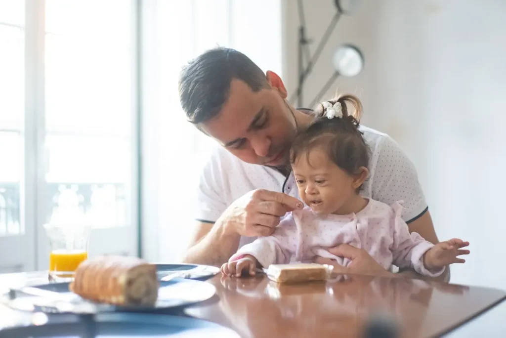 A man wearing a white shirt is leaning over a dark table, gently dabbing the mouth of a toddler girl with Down syndrome after giving her a small piece of food. The bright morning light streams in from a window behind them.