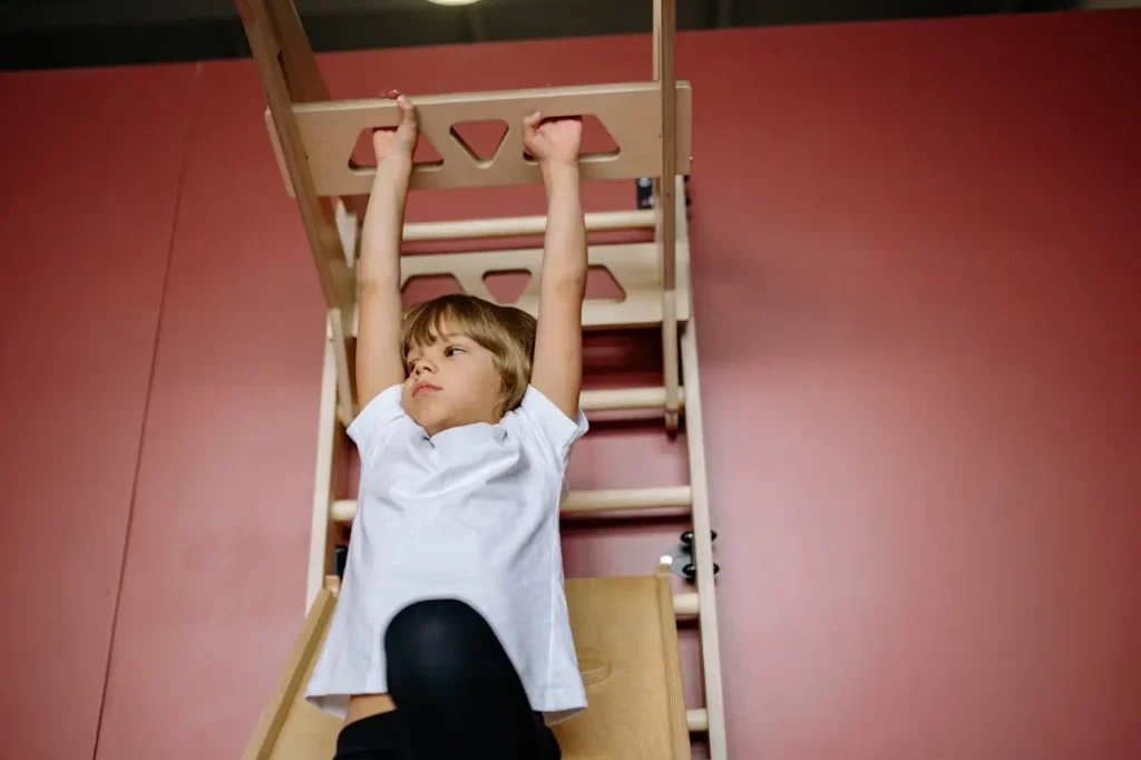 A young boy with light brown hair and a white shirt is hanging from the horizontal ladder (monkey bars) portion of an indoor wooden climbing structure, using an overhead grip. He is actively engaging his arms and core strength.