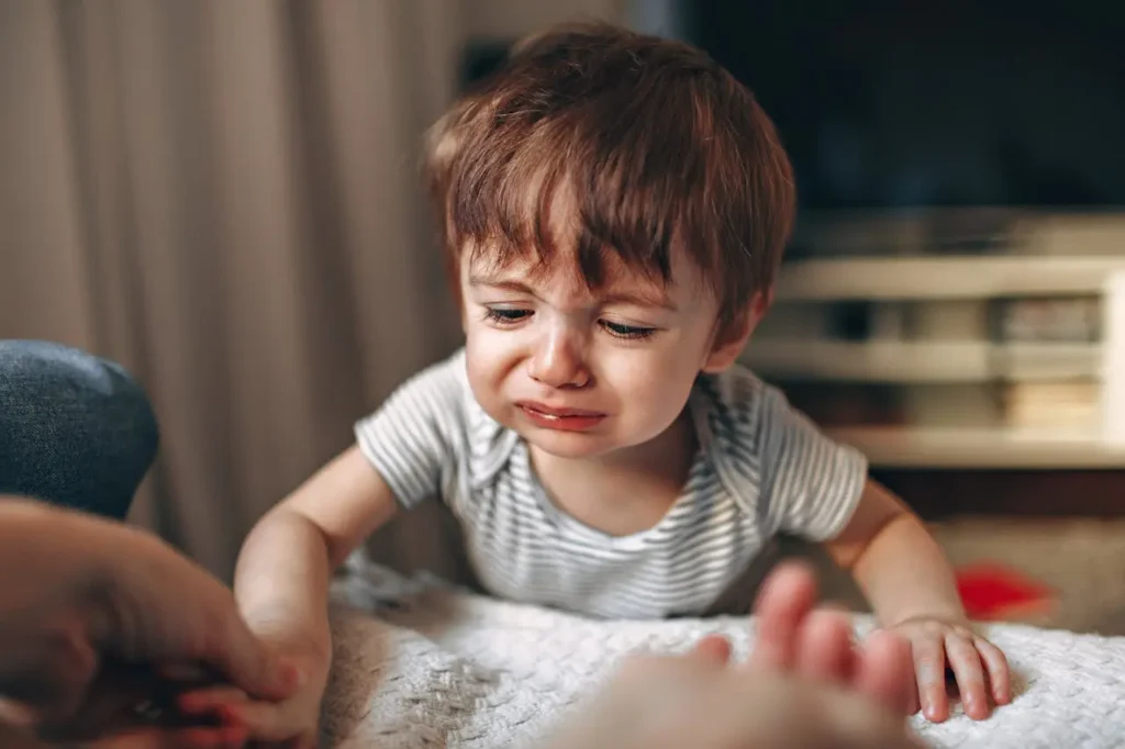 A close-up of a small toddler boy with reddish-brown hair crying intensely, reaching out his hand toward a blurry adult hand for comfort. His face shows clear distress.