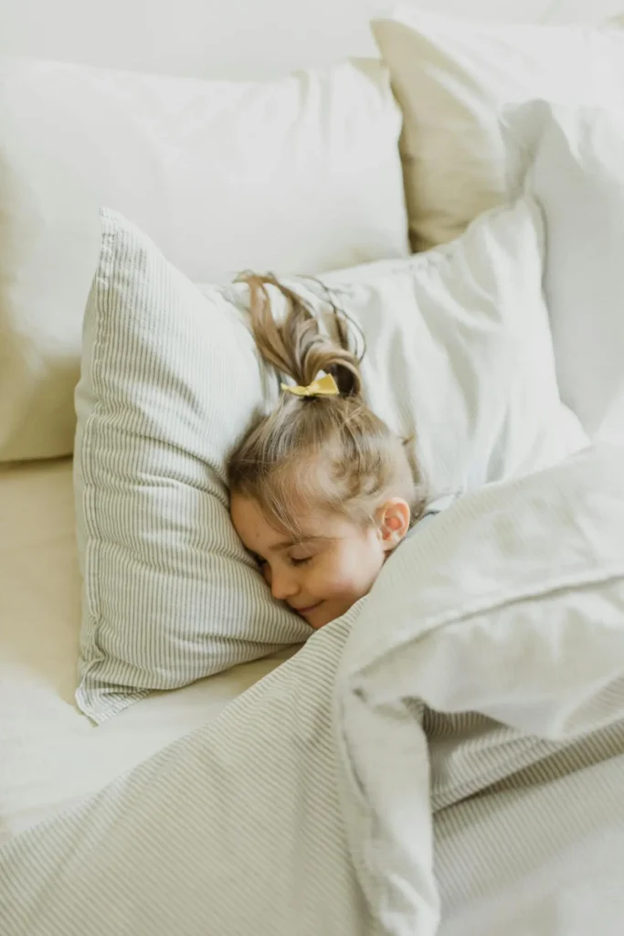 A close-up of a young girl with long brown hair, tied with a yellow bow, sleeping soundly on a large, soft white pillow with faint gray stripes. She is tucked under a matching striped comforter.