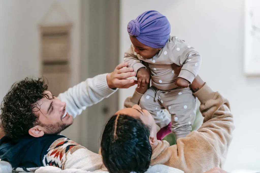 A two-parent family lying on their backs on a white surface, looking up and smiling at their baby who is sitting on the chest of one parent. The baby is wearing a light outfit and a purple head wrap, and the parents are making eye contact and engaging in conversation.