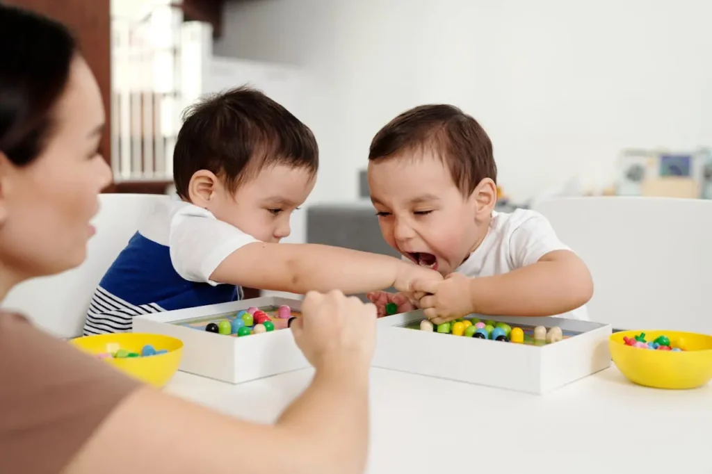 Two twin or young toddler boys are sitting at a table engaged in a conflict, with one boy biting or grasping the arm of the other boy, who is crying out. An adult's hand is visible attempting to intervene, illustrating the need for guidance on How to Handle Toddler Hitting and Biting.