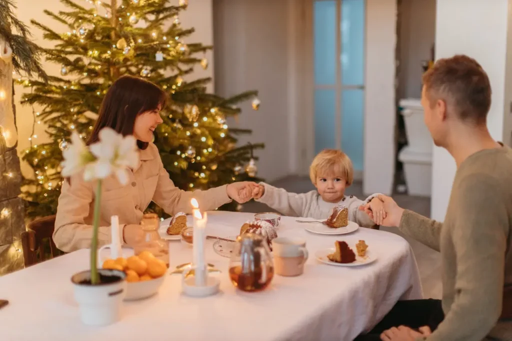 A family of three—a mother, a father, and a toddler boy—sit at a dining table covered with a white cloth, holding hands and smiling while eating slices of cake or dessert. A candle and a decorated Christmas tree are visible in the background.