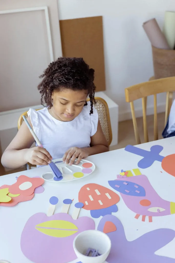 A young girl with curly dark hair is sitting at a white table, intently painting or gluing colorful, geometric paper cutouts of flowers and animals onto a white surface. She is holding a paintbrush and using a palette with purple paint.