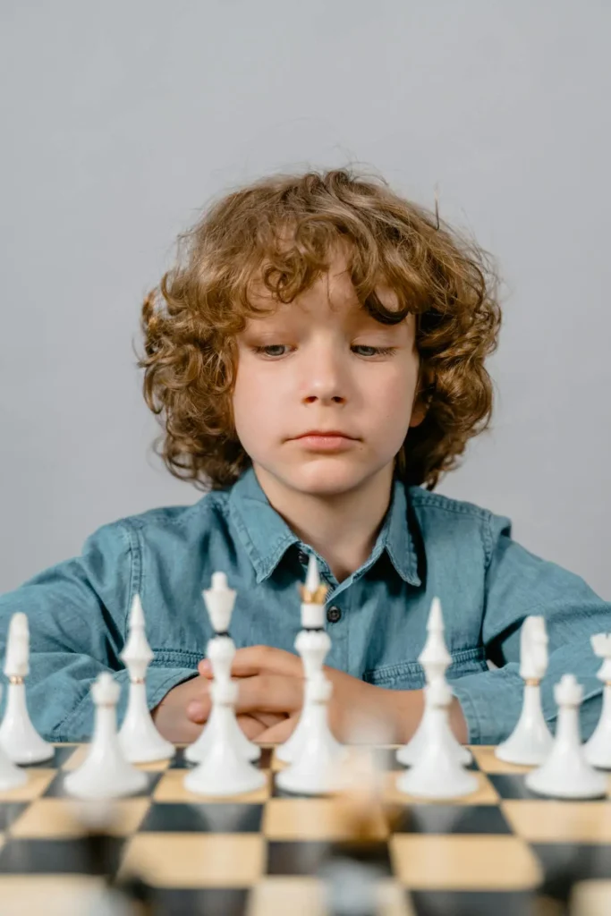 A young boy with curly brown hair and a denim shirt is sitting at a chessboard, intently focusing on the pieces. His serious expression suggests deep thought and the challenge of the game.
