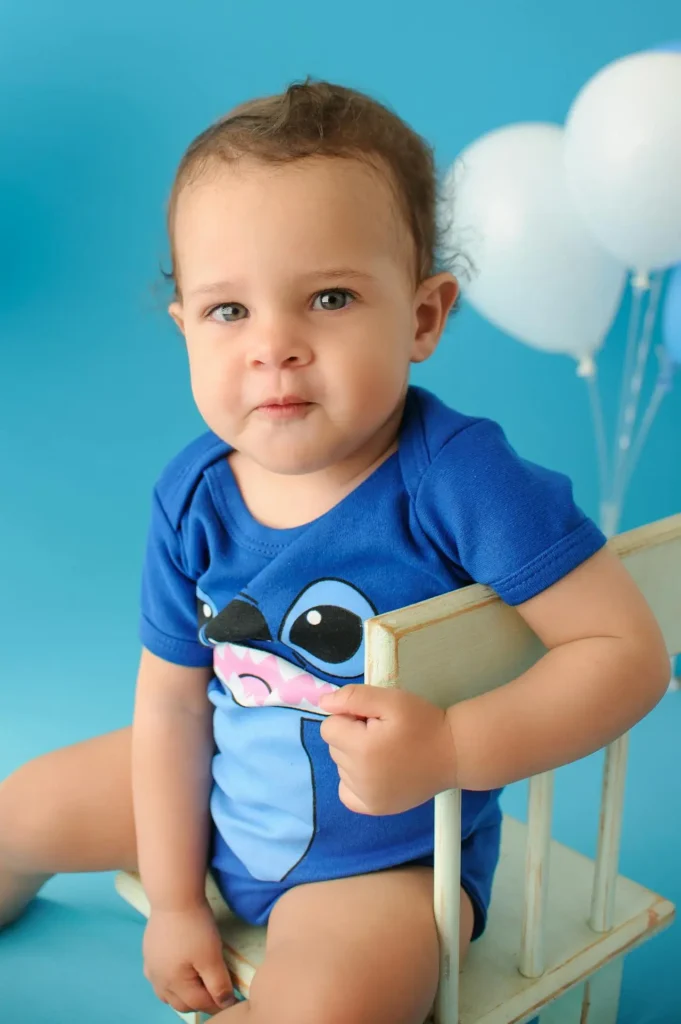 A handsome baby boy with dark curly hair, wearing a blue onesie, is sitting up straight on a small white wooden chair against a bright blue background. His attentive expression suggests listening or an attempt at communication, key to reaching language milestones. White and blue balloons are visible in the background.