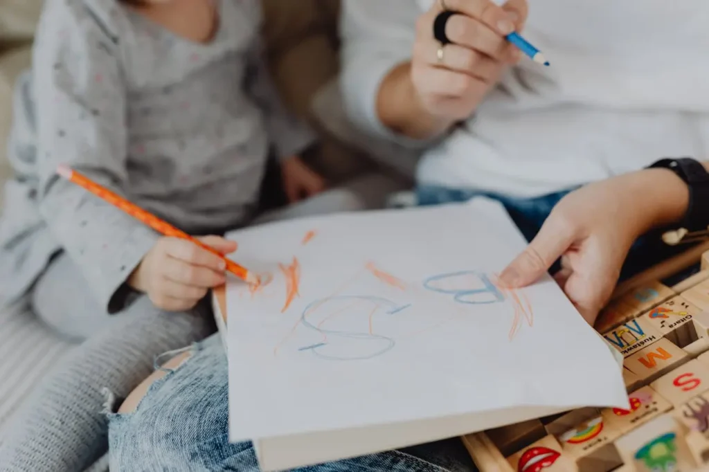 A close-up of a child's hand holding an orange crayon to draw on a piece of paper resting on an adult's lap, while the adult holds a blue crayon and guides the paper. Letters are scribbled on the paper.