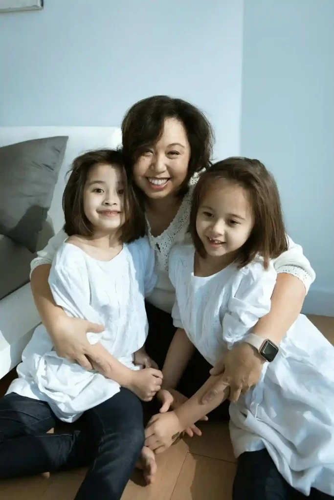 A close-up, warm indoor portrait of a smiling mother sitting with her two young twin daughters who are hugging her. All three are looking directly at the camera, emphasizing connection and happiness.