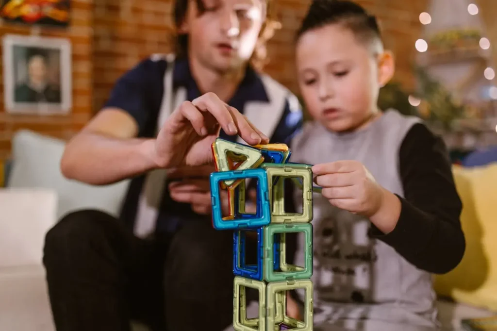 An older boy with dark hair and a younger boy are collaborating on building a tall structure using colorful, square magnetic tiles, demonstrating focused, hands-on play.