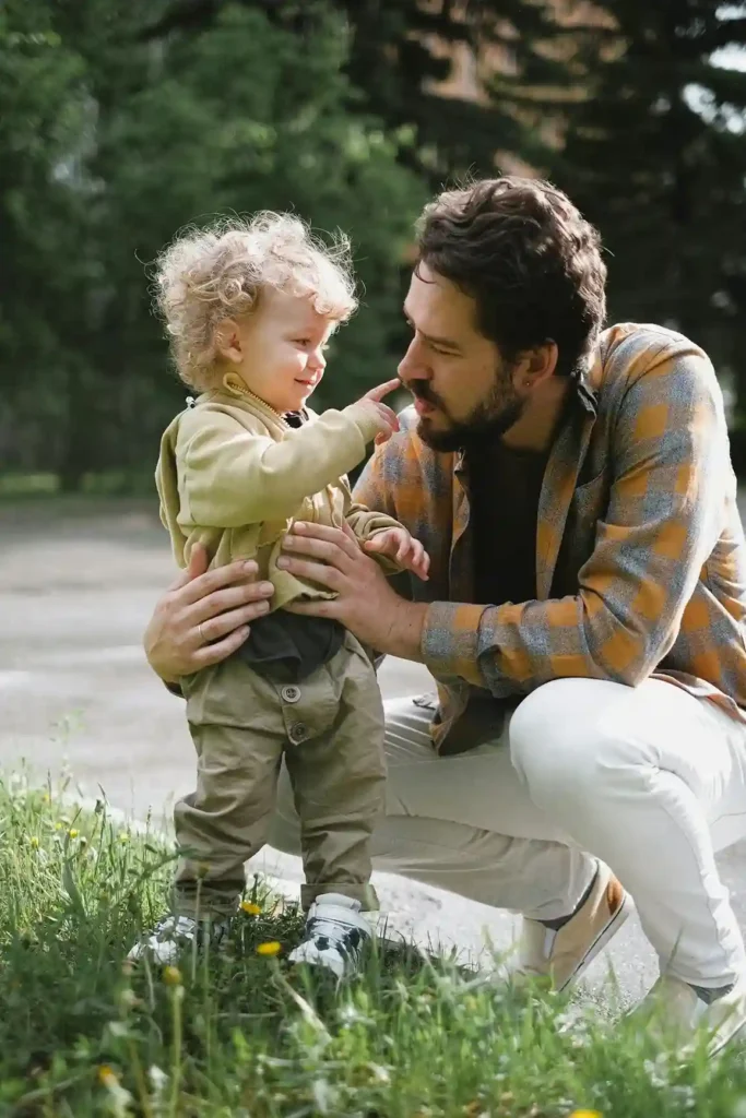 A man with a beard and a plaid shirt is crouching down to interact face-to-face with a toddler with curly blonde hair. The toddler is playfully touching the man's nose, showcasing a positive, engaged parent-child relationship.