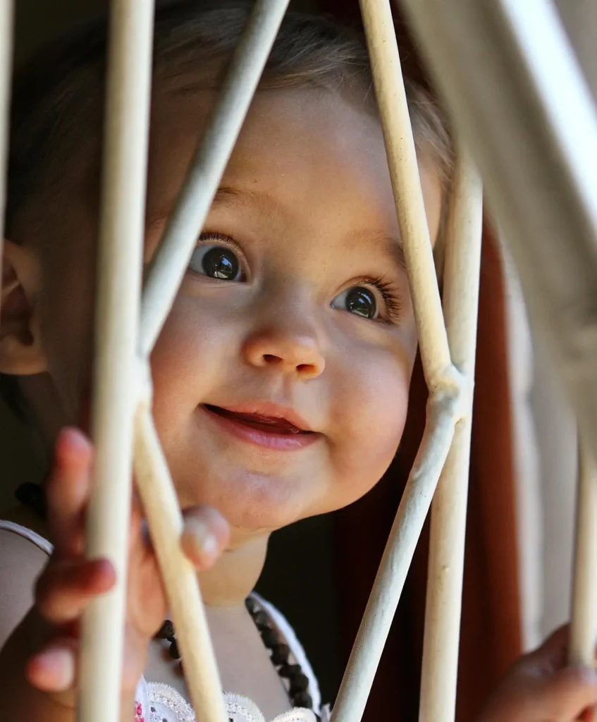 A close-up vertical portrait of a smiling baby or young toddler looking up and gripping the vertical slats of a white crib or safety gate. The child's bright, wide eyes convey curiosity and engagement.