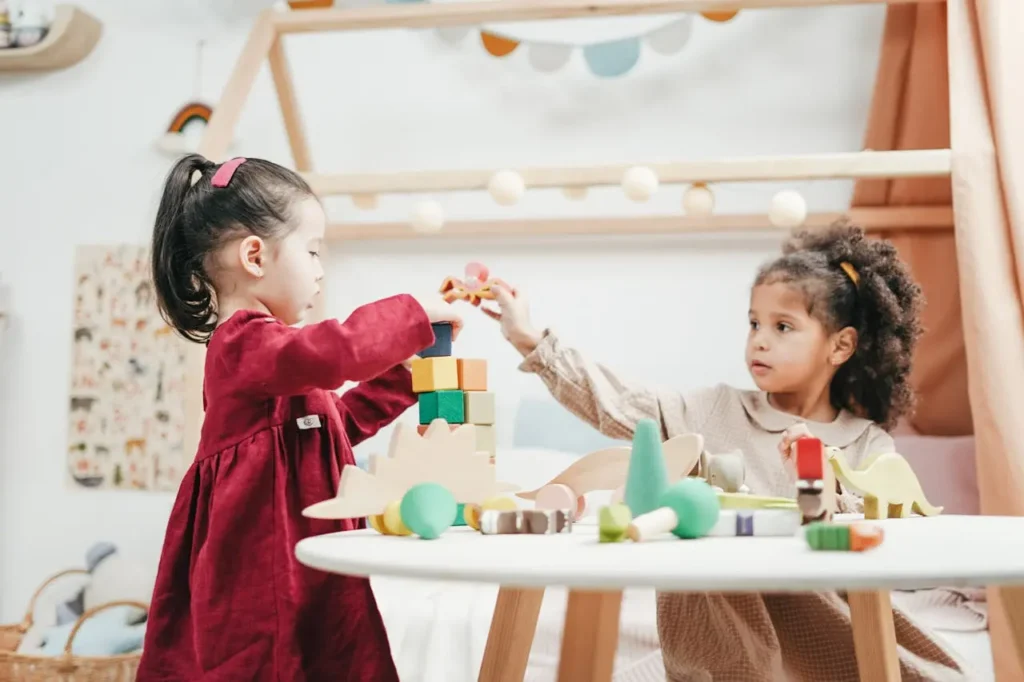 Two young girls, seated near a small wooden table in a play area, are collaborating to build and interact with wooden blocks and small animal figurines. The imaginative play is supported by open-ended-toys-for-toddler-development.