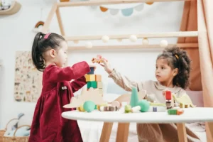 Two young girls, seated near a small wooden table in a play area, are collaborating to build and interact with wooden blocks and small animal figurines. The imaginative play is supported by open-ended-toys-for-toddler-development.