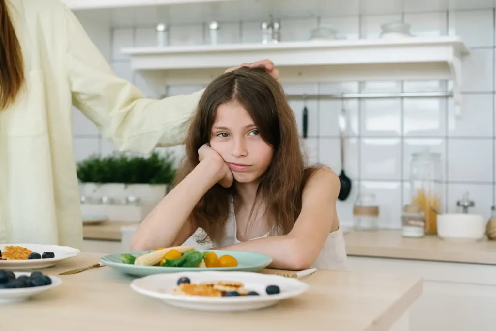A young girl with long brown hair sits with her head propped on her hand at a kitchen table, looking unhappy and bored next to a plate of healthy food (salad/vegetables) that she is refusing to eat. An adult hand is resting reassuringly on her head.