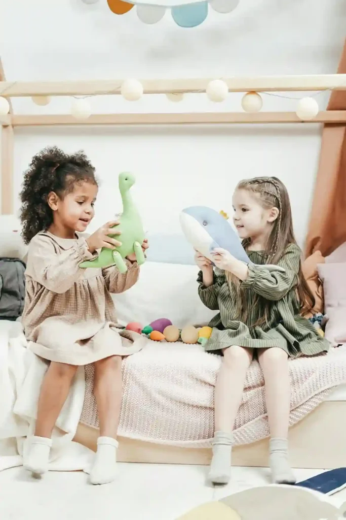 Two preschool-aged girls, one with curly dark hair and one with braided blonde hair, are sitting on a wooden house-frame bed, smiling and holding a green dinosaur and a blue whale stuffed animal, respectively.