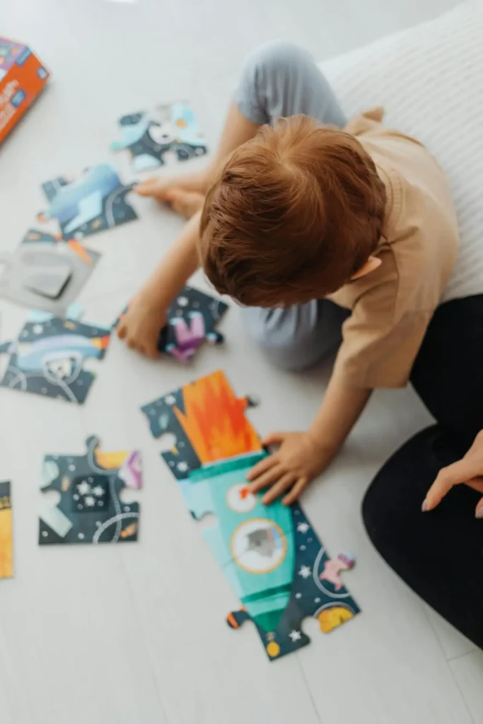 A high-angle shot of a toddler boy with reddish-brown hair sitting on a light floor, concentrating as he uses both hands to connect two large, colorful puzzle pieces depicting a rocket ship. This activity is building problem-solving skills for toddler.
