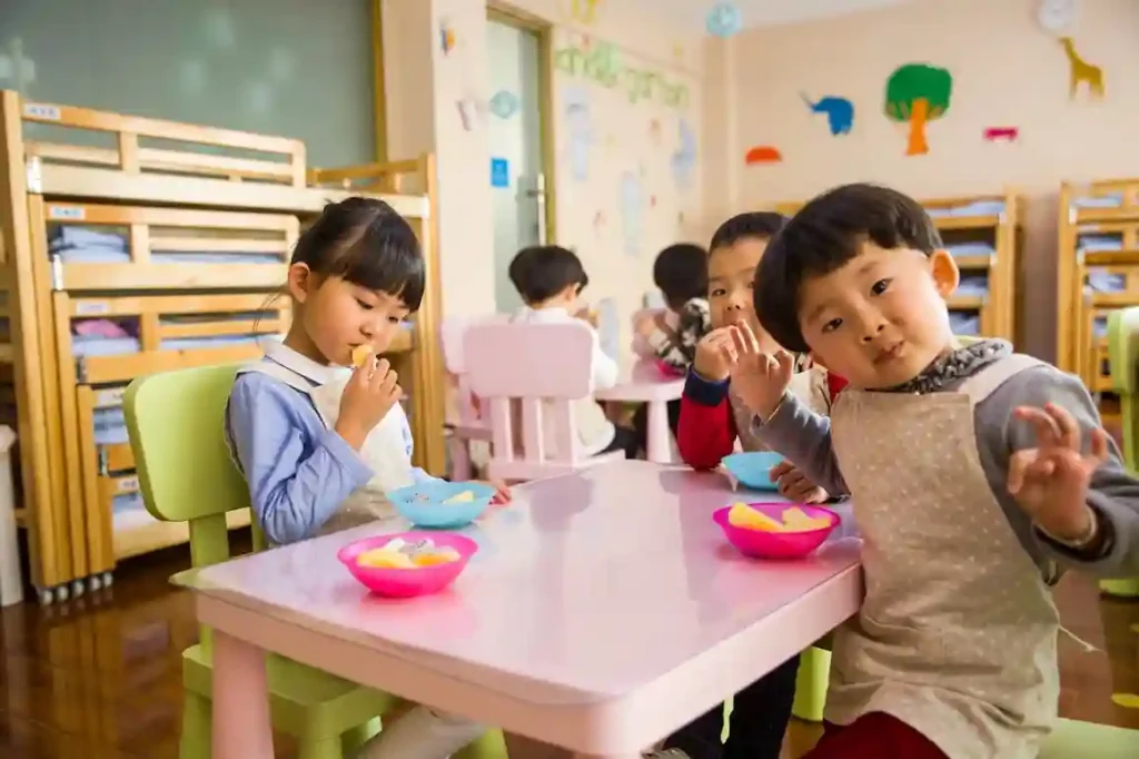 Two Asian children sitting at a small, low pink table in a colorful daycare room, eating fruit from pink and blue bowls. The boy in the foreground is looking at the camera and holding up his hand.